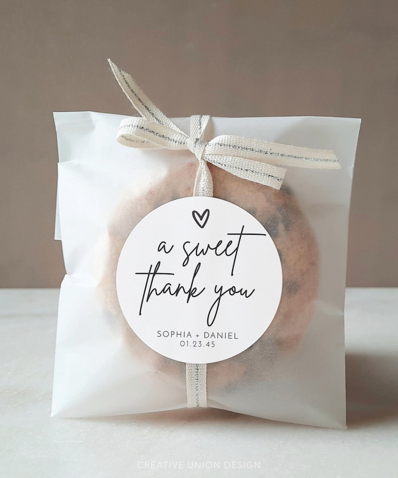 K&ouml;nnte beinhalten: Ein wei&szlig;er Papierbeutel mit einem silbernen Band, das um ihn herum gebunden ist. Der Beutel enth&auml;lt einen Keks mit einem runden wei&szlig;en Aufkleber, auf dem "a sweet thank you" mit einem Herz &uuml;ber dem Text steht. Der Aufkleber sagt auch "Sophia + Daniel 01.23.45"