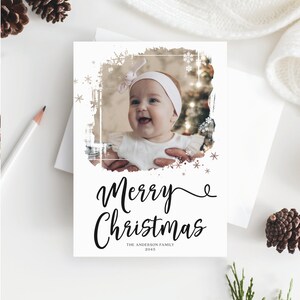 May include: A white Christmas card with a photo of a baby girl wearing a white headband. The card has the words "Merry Christmas" in elegant script, with "The Anderson Family 2015" below. Decorative snowflakes and a distressed border frame the photo.