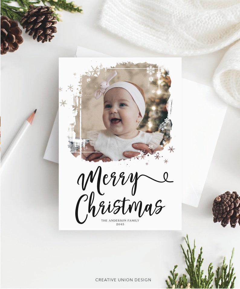 May include: A white Christmas card with a photo of a baby girl wearing a white headband. The card has the words "Merry Christmas" in elegant script, with "The Anderson Family 2015" below. Decorative snowflakes and a distressed border frame the photo.