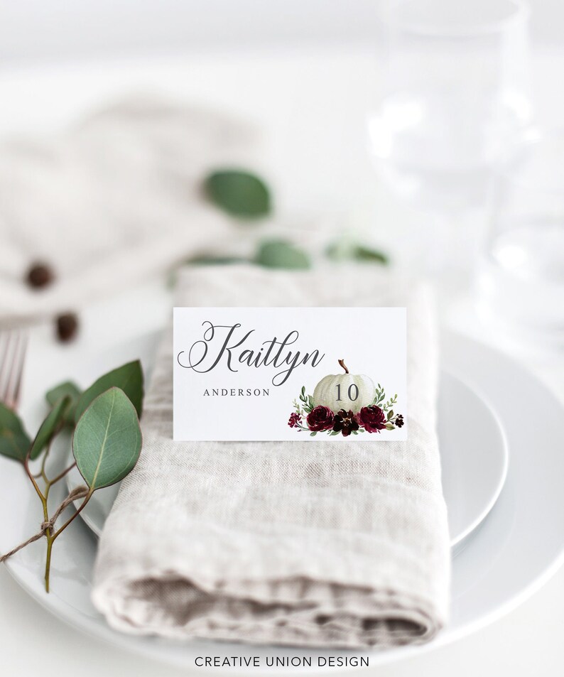 May include: A white place card with the name "Kaitlyn" in elegant script, and the name "Anderson" below. A white pumpkin with the number "10" and floral accents of burgundy and green are on the card. The card rests on a linen napkin.