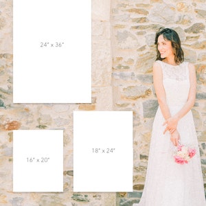 May include: Three white frames with black text indicating different sizes: 24" x 36", 18" x 24", and 16" x 20". The frames are against a stone wall. A woman in a white dress is standing to the right of the frames.