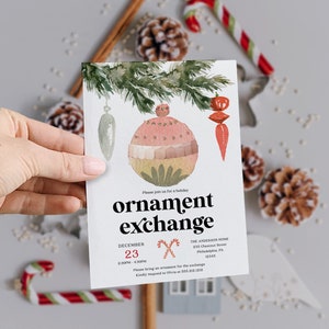 May include: A watercolour invitation for an ornament exchange. The card features a large, decorative ornament, pine needles, and text that reads "ornament exchange" with the date and time. The card is held by a hand.