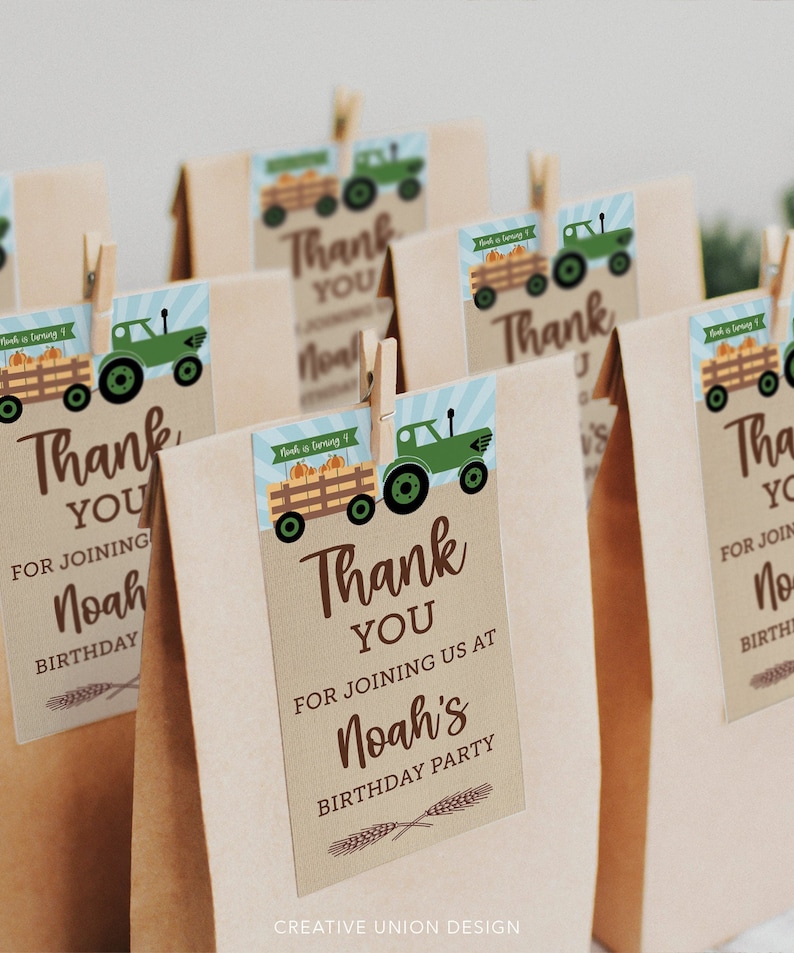 Op de afbeelding: Bruine papieren cadeauzakjes met labels. De labels tonen een groene tractor die een wagen met pompoenen trekt, met de tekst "Bedankt" en "Noah's Verjaardagsfeest". Houten knijpers bevestigen de labels aan de zakjes.