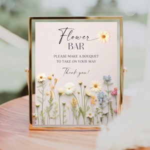 May include: A sign in a gold frame with the text "Flower Bar" and the message "Please make a bouquet to take on your way. Thank you!"  The sign is decorated with dried flowers.