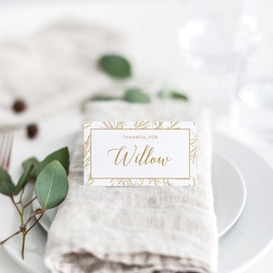Puede incluir: Una tarjeta blanca con un borde dorado y un dise&ntilde;o de hojas doradas. La tarjeta dice "Agradecido por Willow". La tarjeta est&aacute; sobre una servilleta beige en un plato blanco.
