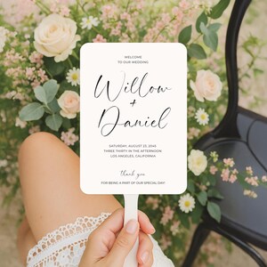 May include: A white wedding fan with the names "Willow + Daniel" in elegant script. The fan includes the wedding date, time, and location. The words "Thank you" are also visible. The background features flowers and greenery.