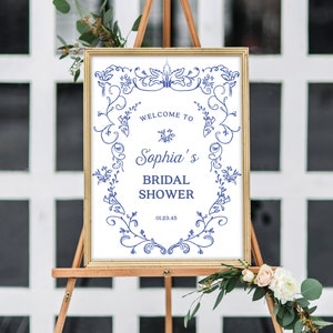 May include: A white sign with a gold frame and blue floral designs. The sign says "Welcome to Sophia's Bridal Shower" and includes the date "01.23.45".
