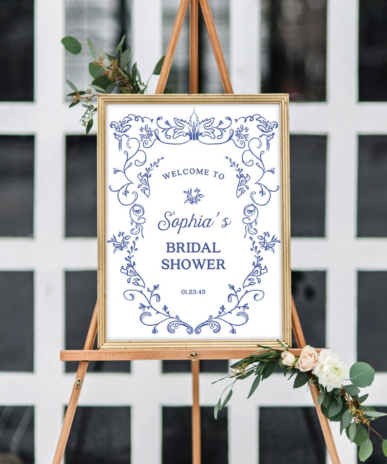 May include: A white sign with a gold frame and blue floral designs. The sign says "Welcome to Sophia's Bridal Shower" and includes the date "01.23.45".