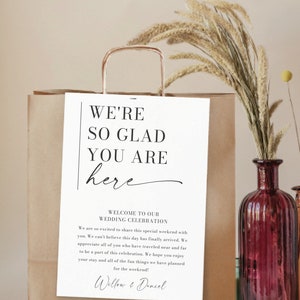 May include: A white sign with black text that reads "We're so glad you are here" and "Welcome to our Wedding Celebration". The sign also includes the names "Willow & Daniel" and the text "Creative Union Design".