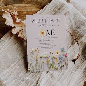 May include: A light purple invitation with a floral design. The text reads "Our Little Wildflower is turning One. Please join us as we celebrate the first birthday of Willow Anderson. Saturday, August 23rd, 2:00pm. The Anderson's, 235 Chestnut Street, Philadelphia, PA. Kindly respond to Jessica at 555.212.1212. Creative Union Design."