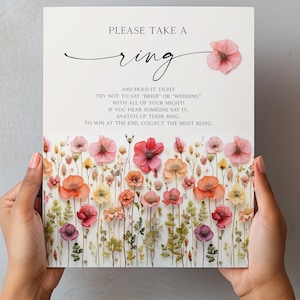 K&ouml;nnte beinhalten: Ein Hochzeitsspielschild mit dem Text "PLEASE TAKE A ring" und Anweisungen. Das Schild ist mit bunten Blumen in Rosa-, Orange- und Gelbt&ouml;nen verziert. Das Schild wird von zwei H&auml;nden gehalten.