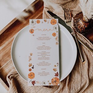 May include: A menu card with a fall theme, featuring watercolor pumpkins and leaves. The menu lists starter, main courses like beef and chicken, and dessert. The card is on a white plate with silverware and a decorative background.