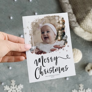 May include: A white Christmas card with a photo of a smiling baby wearing a headband. The card has the words "Merry Christmas" in elegant script, with "The Anderson Family 2045" below. The card is decorated with snowflakes and a distressed border.