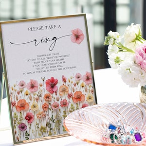 May include: A white sign with a gold frame and pink flowers printed on it. The sign says "Please take a ring and hold it tight. Try not to say "bride" or "wedding" with all of your might! If you hear someone say it, snatch up their ring. To win at the end, collect the most bling."