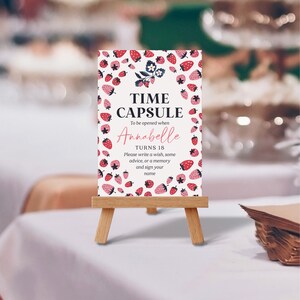 May include: A white sign with a strawberry pattern and the text "TIME CAPSULE To be opened when Annabelle TURNS 18 Please write a wish, some advice, or a memory and sign your name". The sign is on a small wooden easel.