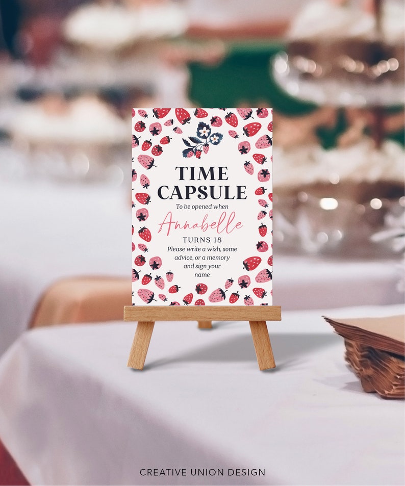 May include: A white sign with a strawberry pattern and the text "TIME CAPSULE To be opened when Annabelle TURNS 18 Please write a wish, some advice, or a memory and sign your name". The sign is on a small wooden easel.