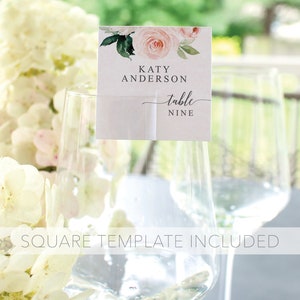 May include: A white card with pink and green watercolor floral design. The card says "Katy Anderson table nine". The card is placed on a clear glass.