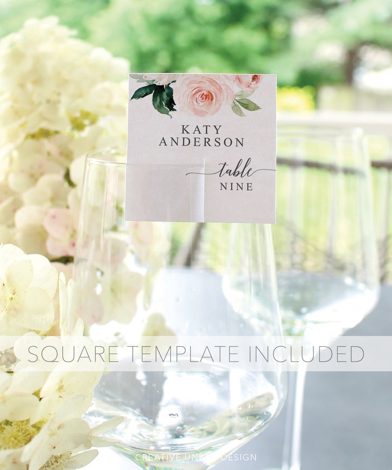 May include: A white card with pink and green watercolor floral design. The card says "Katy Anderson table nine". The card is placed on a clear glass.
