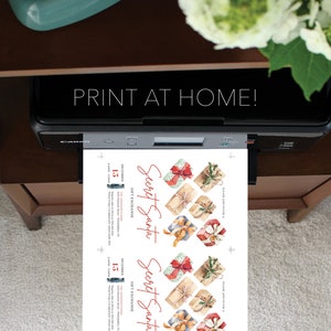 May include: A black printer outputting a white sheet of paper with two identical Secret Santa gift exchange invitations. The invitations feature watercolor illustrations of wrapped presents with red, blue, and tan ribbons and text detailing a December 15 event.