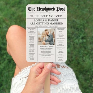 May include: A wedding program fan with a newspaper design. The headline reads "The Newlywed Post" and the subheading reads "The Best Day Ever." The program lists the wedding party, ceremony details, and a message to family and friends. A photo of the couple is included.
