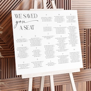 Puede incluir: Un letrero blanco sobre un caballete de madera con las palabras "WE SAVED YOU A SEAT" en escritura elegante. El letrero enumera los nombres alfabéticamente, probablemente para una boda o evento, con un fondo de madera decorativo.