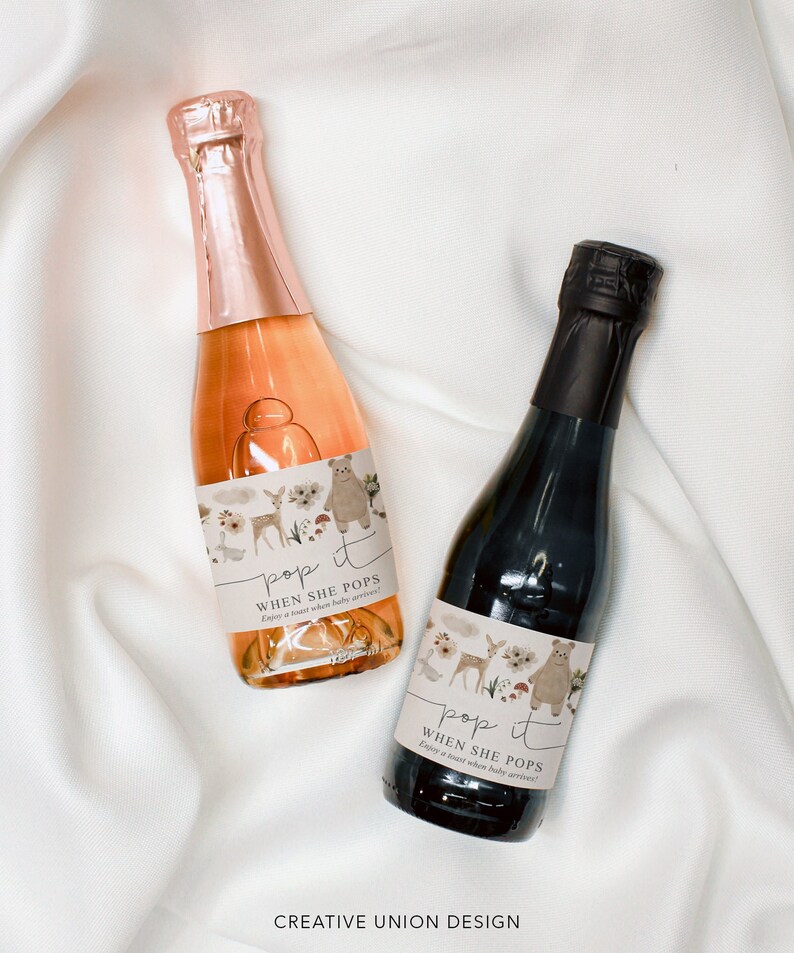 Puede incluir: Dos mini botellas de vino espumoso con etiquetas que dicen "pop it when she pops" y presentan un tema de animales del bosque con un ciervo, un oso y setas. Las etiquetas son beige con acentos marrones y verdes.