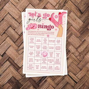 May include: A pink and white bingo card with the text "Let's go girls Bingo" at the top. The card has a variety of squares with different prompts for a bridal shower game. The card features a pink disco ball in the center.
