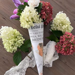 以下が含まれることがあります： 女性の写真と新聞風のデザインが施された白い紙の円錐形。円錐形には、白、ピンク、紫の花のブーケが入っています。円錐形には、「Bridal Party」と「質問する番です。ソフィア、私のブライズメイドになってくれますか？」という文字が書かれています。円錐形は白いレースのリボンで結ばれており、下部に「Ashley」という名前が書かれています。