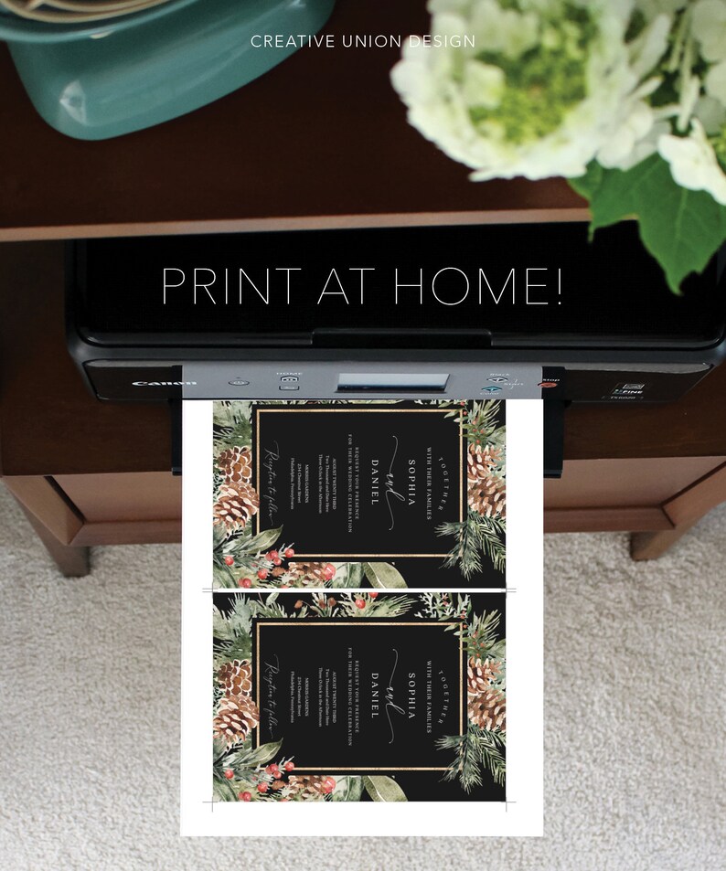 May include: A printer is shown printing two wedding invitations. The invitations have a black background with gold borders and floral and pinecone accents. The text "PRINT AT HOME!" is displayed above the printer.
