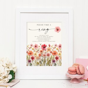 May include: A white framed sign with a floral background and the text "PLEASE TAKE A ring AND HOLD IT TIGHT TRY NOT TO SAY "BRIDE" OR "WEDDING" WITH ALL OF YOUR MIGHTY IF YOU HEAR SOMEONE SAY IT, SNATCH UP THEIR RING TO WIN AT THE END, COLLECT THE MOST BLING" and the text "CREATIVE UNION DESIGN" at the bottom.