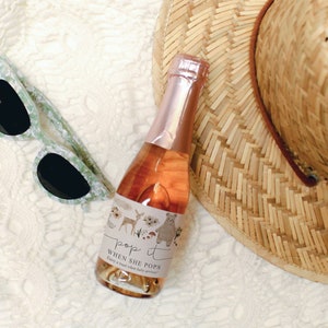 Puede incluir: Una peque&ntilde;a botella de champ&aacute;n rosado con una etiqueta que dice "Pop it when she pops! Enjoy a toast when baby arrives!" La botella est&aacute; sobre una superficie blanca con un sombrero de paja y gafas de sol.