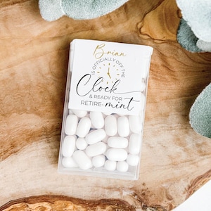 May include: A clear plastic box filled with white mints, with a label that reads "Brian is officially off the clock & ready for retire-mint." The label has gold and black text. The box is on a wooden surface.