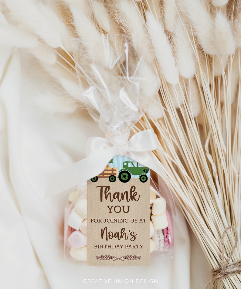 Op de afbeelding: Een transparante cellofaan zak, vastgebonden met een wit lint, gevuld met marshmallows en een label. Op het label staat "Thank You for joining us at Noah's Birthday Party" met een groene tractorafbeelding. Gedroogde tarwestengels staan op de achtergrond.