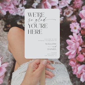 May include: A rectangular wedding sign with rounded corners, held by a person. The sign reads "WE'RE so glad YOU'RE HERE" in elegant black script. Below, it states "WELCOME TO THE WEDDING CELEBRATION OF WILLOW and DANIEL". Pink flowers are in the background.