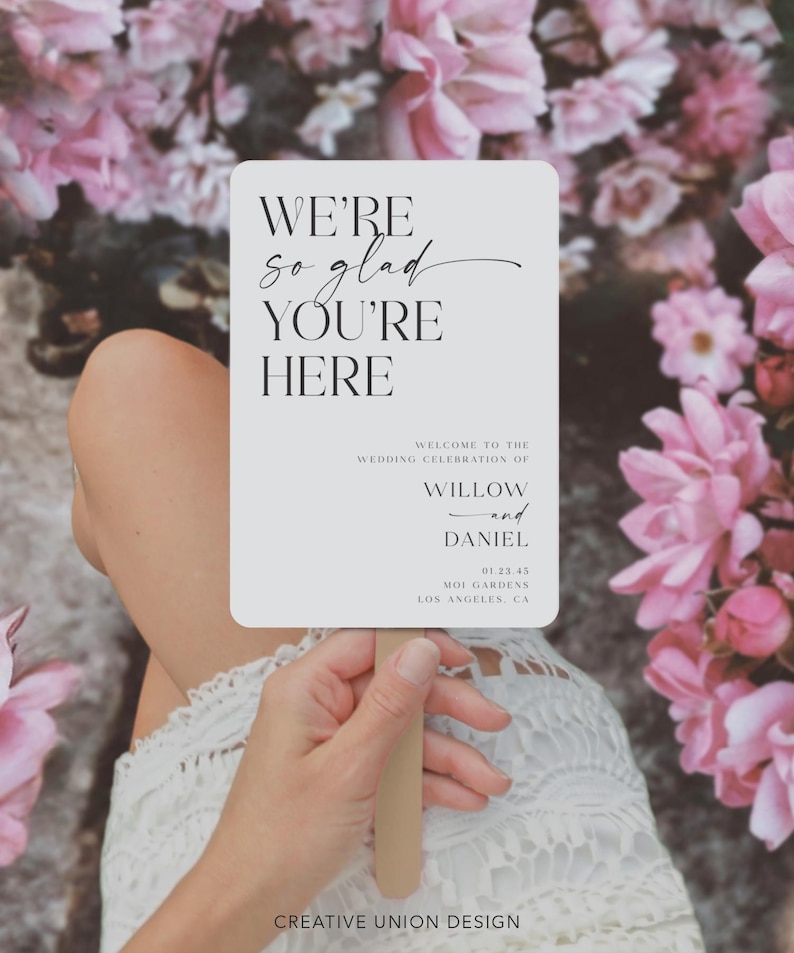 May include: A rectangular wedding sign with rounded corners, held by a person. The sign reads "WE'RE so glad YOU'RE HERE" in elegant black script. Below, it states "WELCOME TO THE WEDDING CELEBRATION OF WILLOW and DANIEL". Pink flowers are in the background.