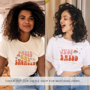 May include: Two women wearing white t-shirts with different designs. The first shirt says "Dazed and Engaged" in a retro font with a mushroom and diamond graphic. The second shirt says "Just Dazed" in a retro font with a mushroom graphic.