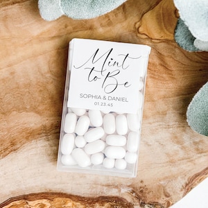 May include: A clear plastic box filled with white mints, with a white label that reads "Mint to Be" in elegant script, along with the names "Sophia & Daniel" and a date. The box is on a wooden surface.