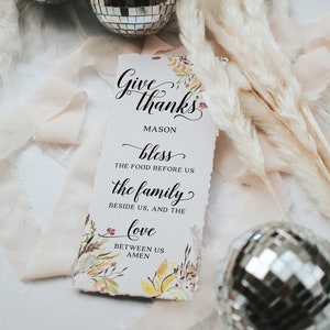 May include: A rectangular paper sign with the words "Give thanks" in elegant script, with floral accents. The sign includes the text "bless the food before us, the family beside us, and the love between us." Two disco balls and decorative dried plants are in the background.