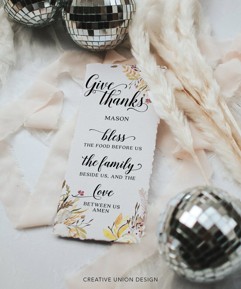 May include: A rectangular paper sign with the words "Give thanks" in elegant script, with floral accents. The sign includes the text "bless the food before us, the family beside us, and the love between us." Two disco balls and decorative dried plants are in the background.