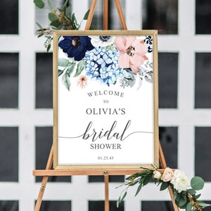 May include: A rectangular sign in a gold frame featuring a floral arrangement of navy, light blue, and peach flowers at the top. The text reads: WELCOME TO OLIVIA'S bridal SHOWER 01.23.45. The sign is displayed on a wooden easel with greenery.