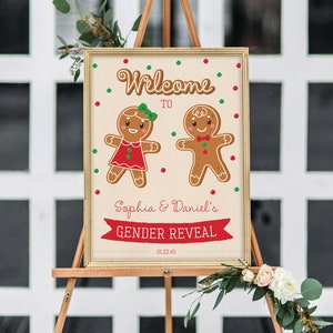 May include: A framed sign on an easel announces a gender reveal for Sophia & Daniel. The sign features gingerbread people and the words "Welcome to" above the names. Red and green dots and a red banner that says "GENDER REVEAL" decorate the sign.