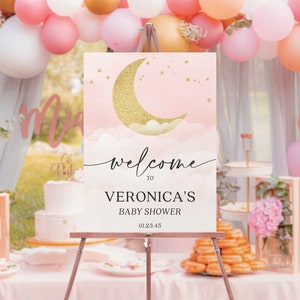 May include: A rectangular welcome sign on a metal easel featuring a gold glitter crescent moon, white clouds, and gold stars on a soft pink background. The text reads: welcome TO VERONICA'S BABY SHOWER 01.23.45 in black lettering.