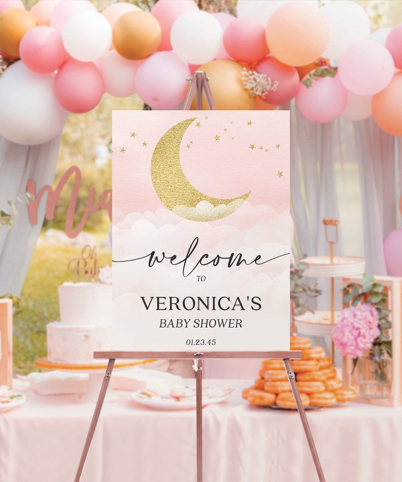 May include: A rectangular welcome sign on a metal easel featuring a gold glitter crescent moon, white clouds, and gold stars on a soft pink background. The text reads: welcome TO VERONICA'S BABY SHOWER 01.23.45 in black lettering.