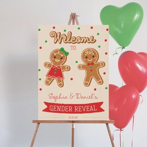 May include: A gender reveal sign on an easel features gingerbread people and the text "Welcome to Sophia & Daniel's Gender Reveal." Red and green heart-shaped balloons are in the background. The sign is decorated with red and green dots.