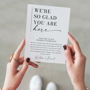 May include: A white card with black text that reads "We're so glad you are here" and "Welcome to our Wedding Celebration". The card also includes a message welcoming guests to the wedding celebration and a signature that reads "Willow & Daniel".