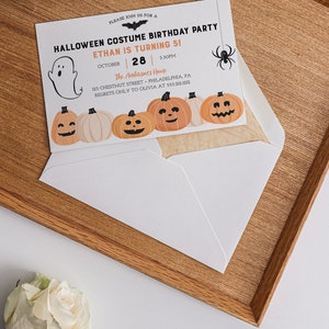 以下が含まれることがあります： 「HALLOWEEN COSTUME BIRTHDAY PARTY」と「ETHAN IS TURNING 5！」の文字が入った、ハロウィーンをテーマにした誕生日パーティーの招待状。カボチャ、幽霊、コウモリ、クモのイラストが特徴です。招待状は白い封筒の中に入っています。