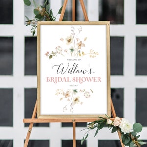May include: A framed sign on a wooden easel announces "Welcome to Willow's Bridal Shower" in elegant script and a soft pink hue. The sign is adorned with floral illustrations in muted tones, with the date 01.23.45. Decorative greenery and flowers frame the sign.