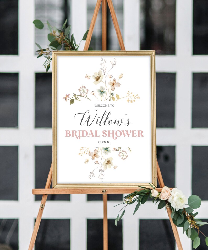 May include: A framed sign on a wooden easel announces "Welcome to Willow's Bridal Shower" in elegant script and a soft pink hue. The sign is adorned with floral illustrations in muted tones, with the date 01.23.45. Decorative greenery and flowers frame the sign.