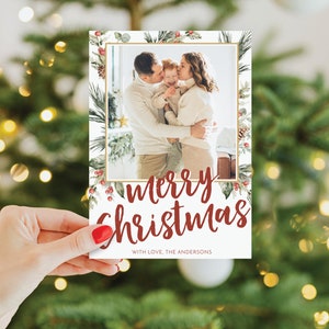 May include: A Christmas card held by a hand, featuring a family photo within a gold frame. The card has a festive design with the words "Merry Christmas" in red script, surrounded by watercolor greenery and berries. The text "WITH LOVE, THE ANDERSONS" is also present.