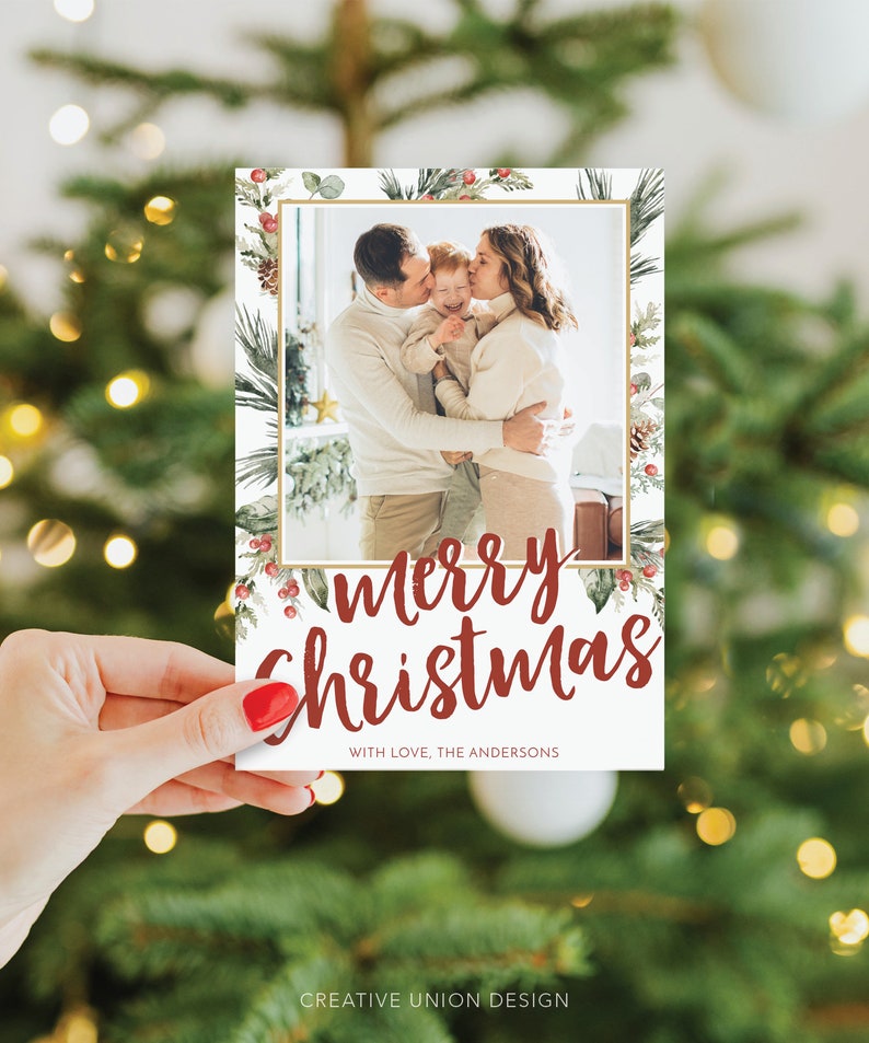 May include: A Christmas card held by a hand, featuring a family photo within a gold frame. The card has a festive design with the words "Merry Christmas" in red script, surrounded by watercolor greenery and berries. The text "WITH LOVE, THE ANDERSONS" is also present.